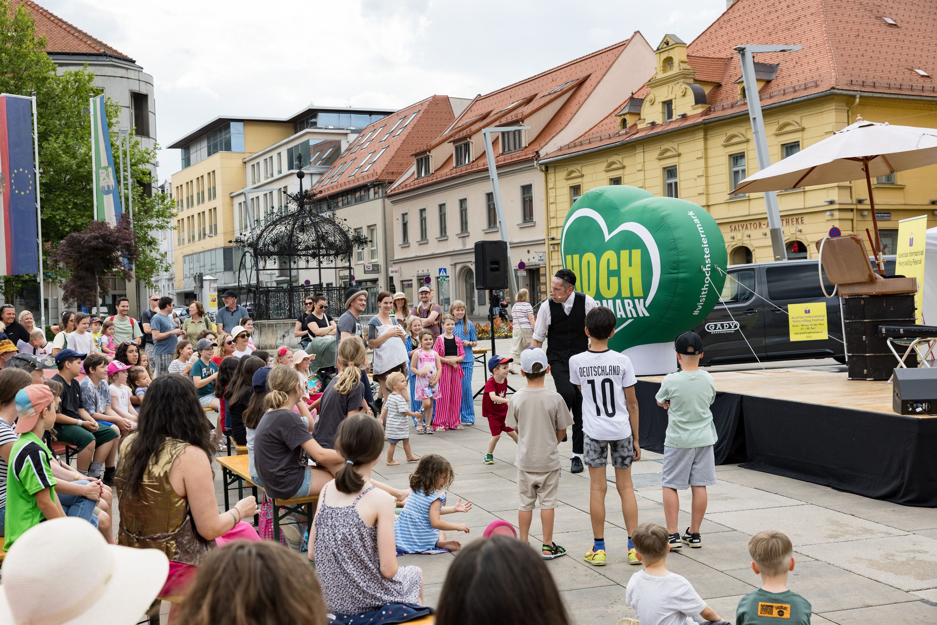 Eine Menschenmenge, einschließlich Kinder, versammelt sich auf einer Straße vor einer Bühne. Ein Mann mit Mikrofon und grünem Ballon steht auf der Bühne.