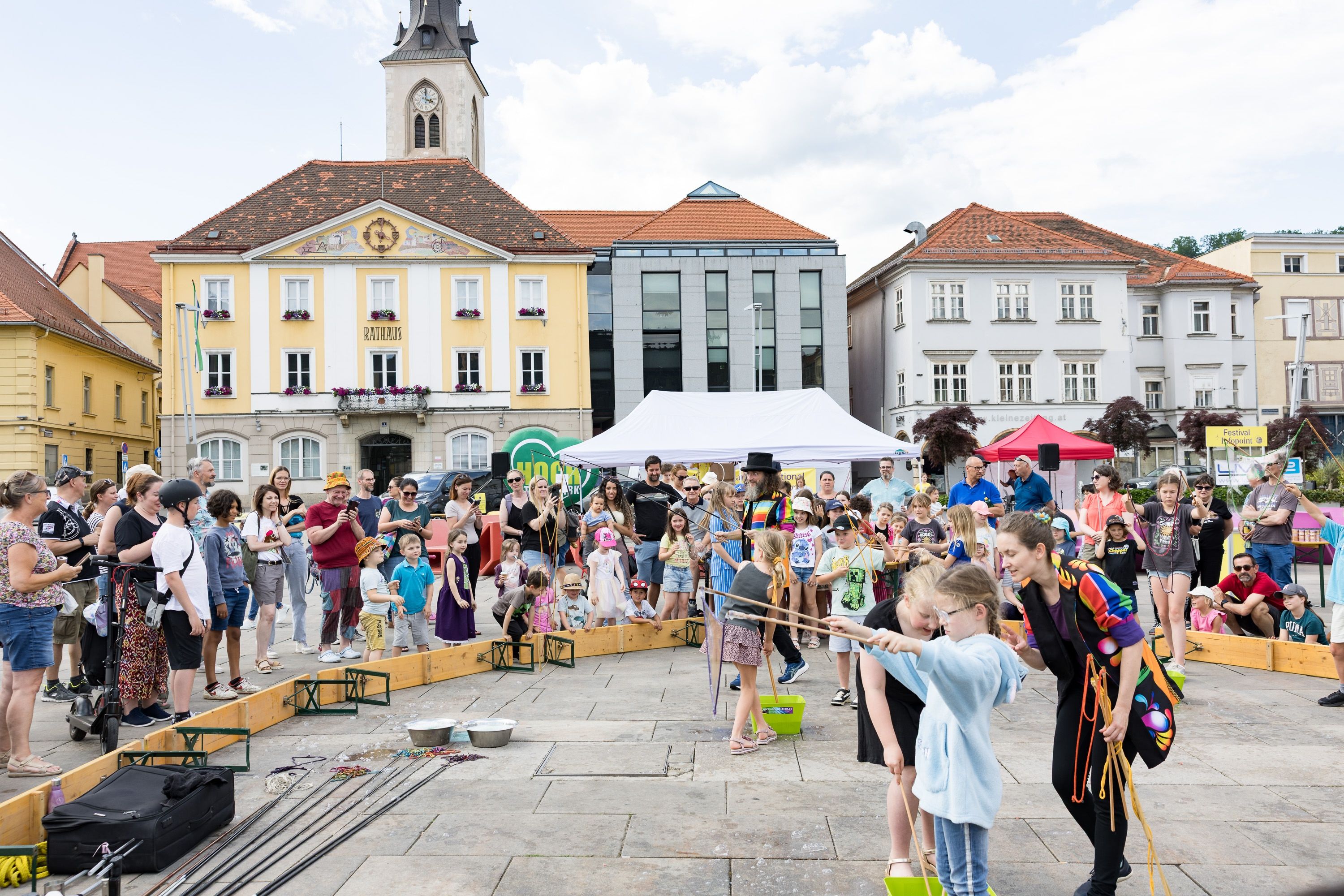 Eine öffentliche Veranstaltung findet auf einem Stadtplatz statt. Mehrere Kinder halten Holzstäbe und führen einen choreografierten Tanz auf. Zuschauer umgeben den Platz. Ein Zelt und ein Zelt mit rotem Dach sind im Hintergrund. Gebäude säumen den Platz.