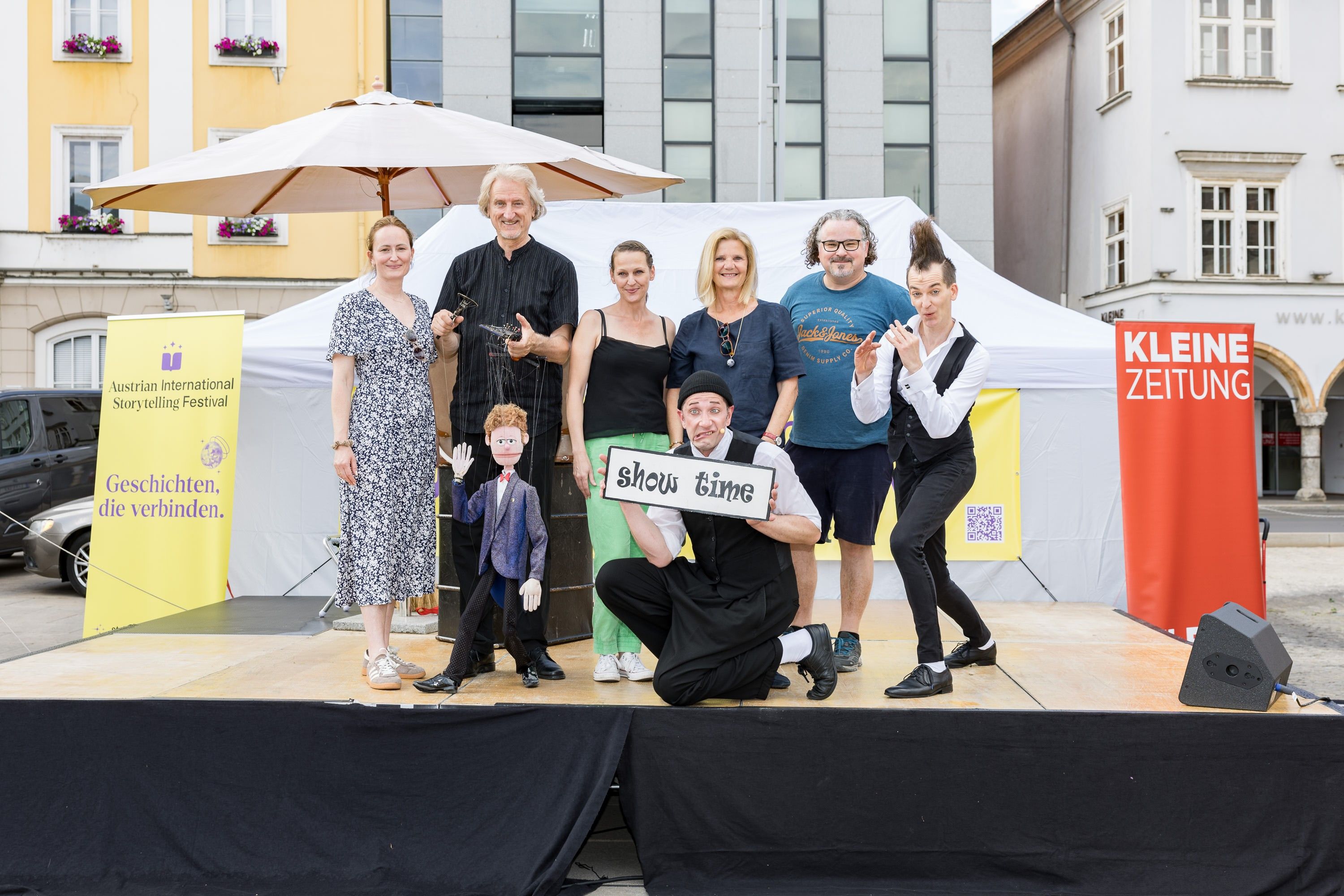 Eine Gruppe von Menschen, darunter ein Mann, der eine Marionette hält, posiert für ein Foto auf der Bühne. Sie lächeln und ein Mann hält ein Schild mit der Aufschrift Show Time.