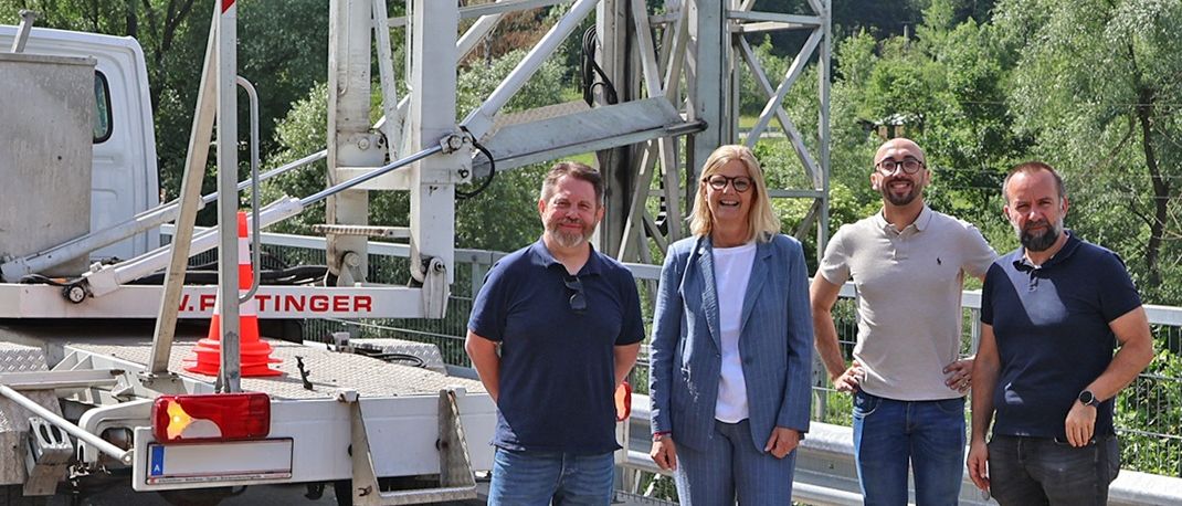 Vier Personen stehen vor einer Brücke mit einem Fahrzeug dahinter. Eine Metallstruktur mit einem Schild und Logo ist am Fahrzeug montiert.