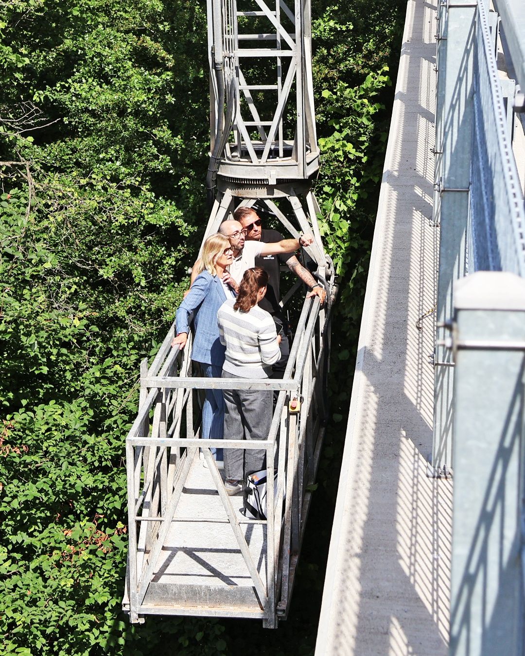 Fünf Personen stehen auf einer Brücke und schauen nach unten. Sie tragen Freizeitkleidung. Die Brücke ist von grünen Pflanzen umgeben.