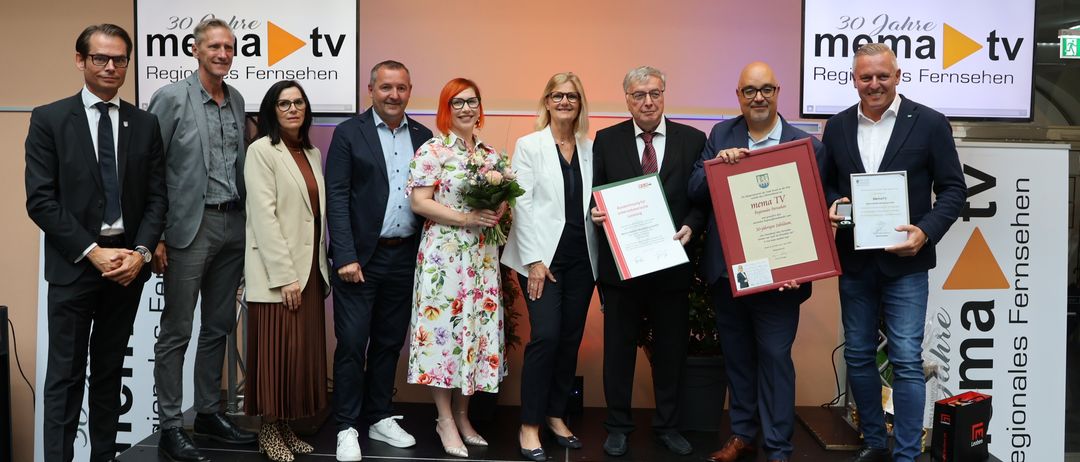 Eine Gruppe von Menschen steht auf einer Bühne und hält Urkunden in der Hand. Eine Frau in der Mitte hält Blumen und eine Urkunde. Hinter ihnen befinden sich zwei Banner mit den Worten 'la tv' und '30 Jahre'. Sie alle lächeln und posieren für ein Foto.