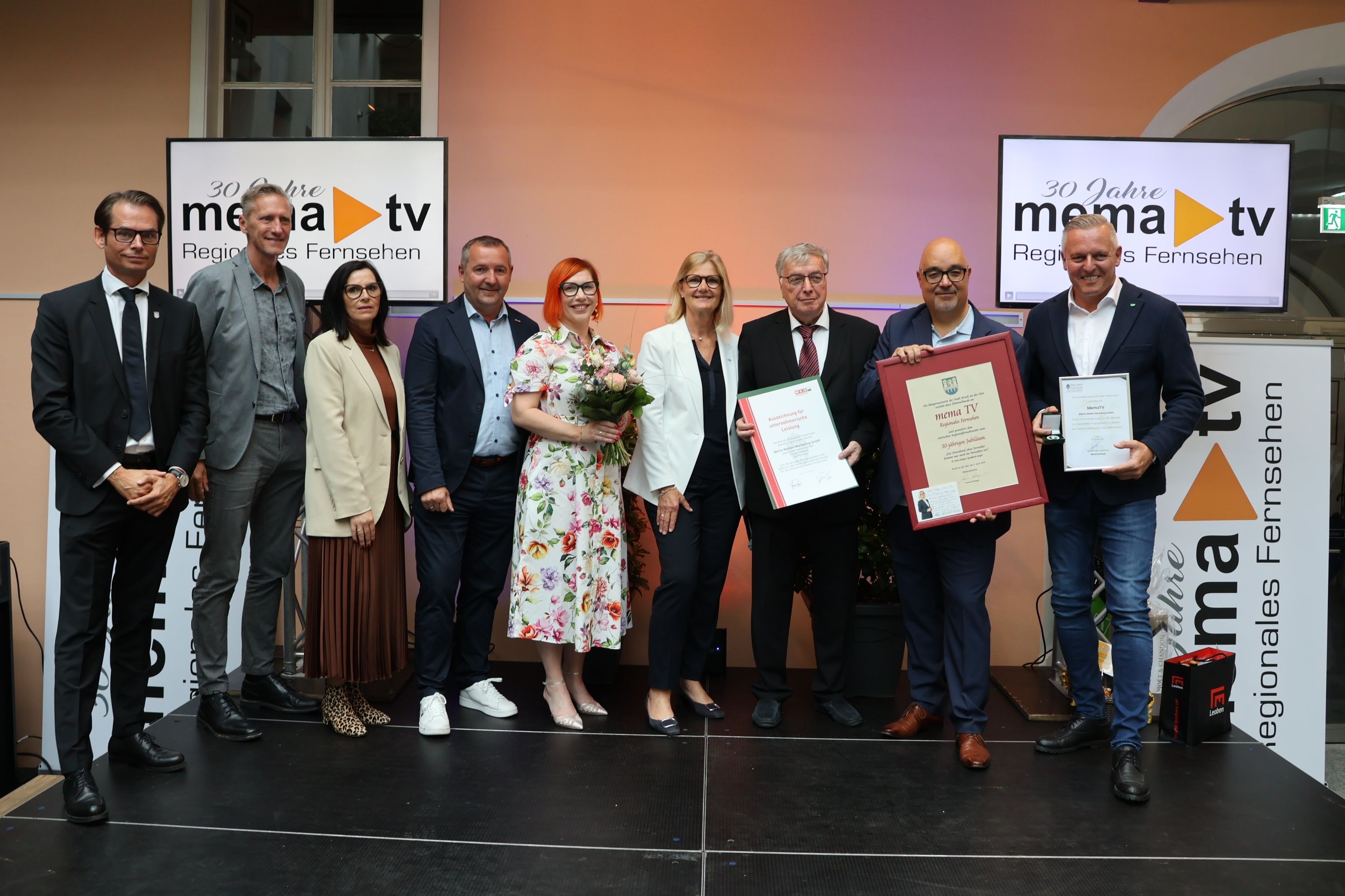 Mehrere Personen, darunter Männer und Frauen, posieren für ein Foto auf der Bühne mit Urkunden und Blumen. Sie sind alle formell gekleidet. Dahinter befindet sich eine Wand mit einem Schild.
