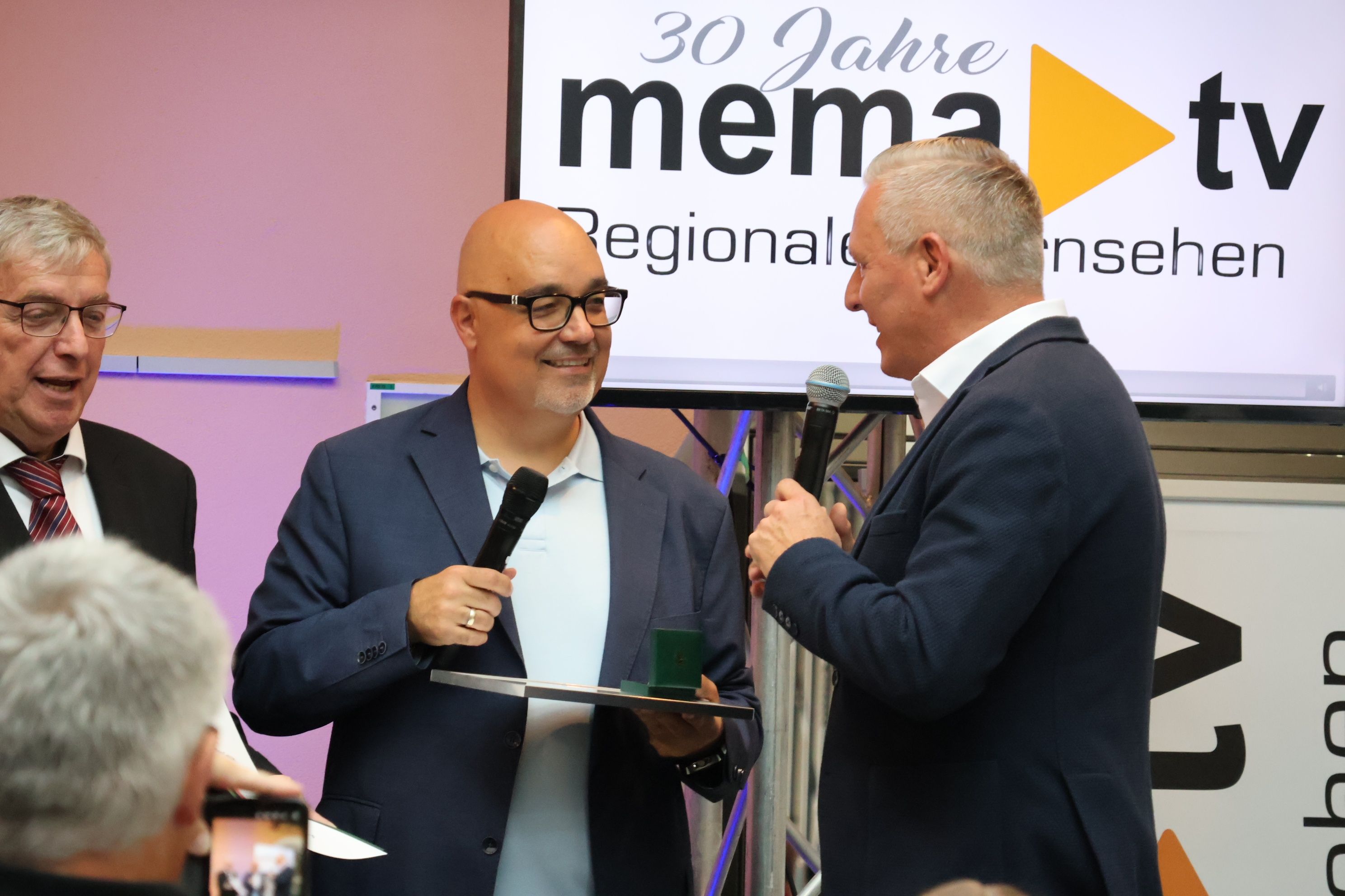 Zwei Männer in Anzügen halten Mikrofone und lächeln bei einer Preisverleihung. Der Mann auf der linken Seite hält eine grüne Schachtel und eine Plakette. Ein Banner mit 30 Jahre und einem gelben Pfeil ist hinter ihnen.