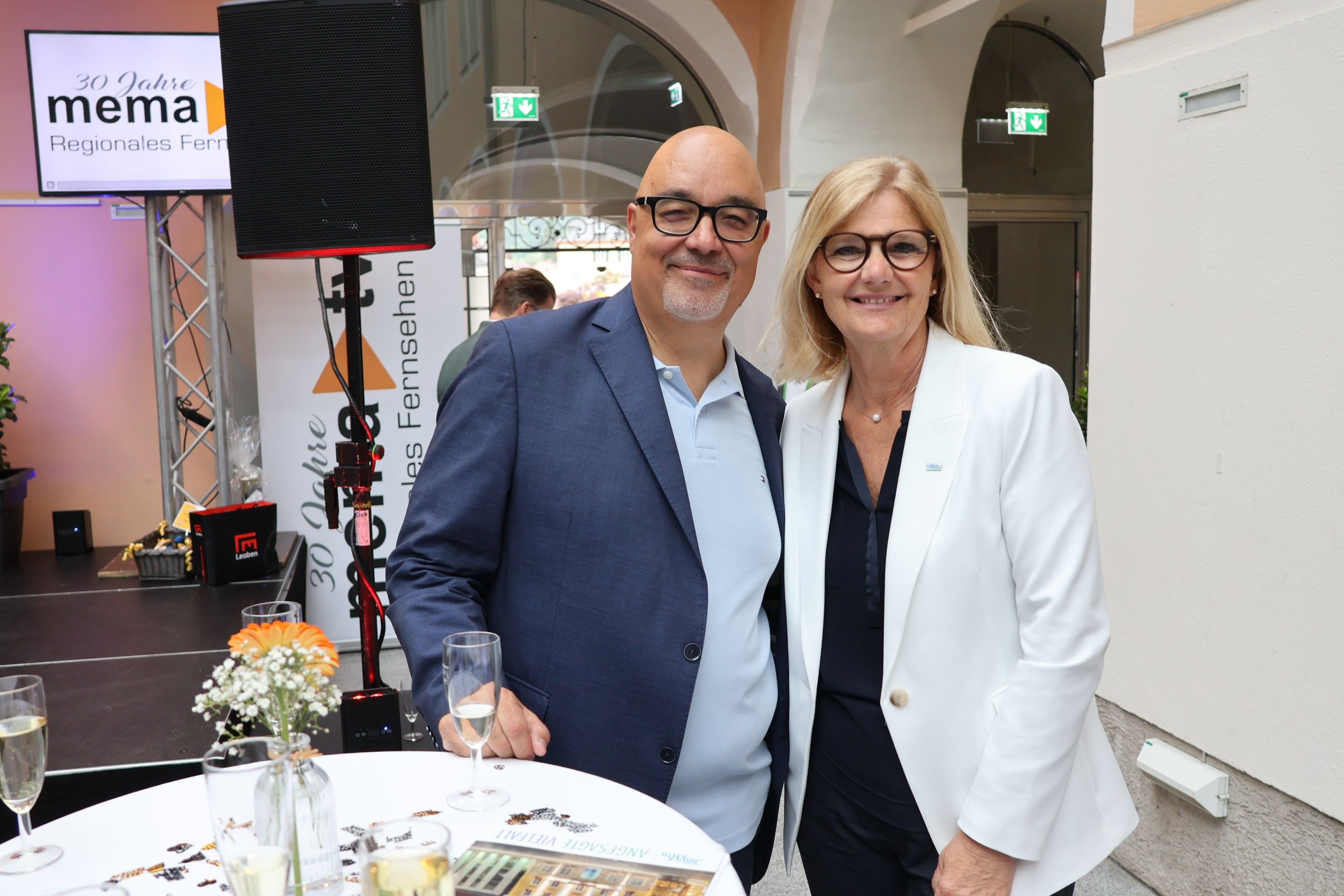 Ein Mann und eine Frau lächeln für ein Foto in einem Raum. Sie stehen neben einem Tisch mit Weingläsern und Blumen. Hinter ihnen steht ein Lautsprecher und ein Schild für ein Unternehmen.