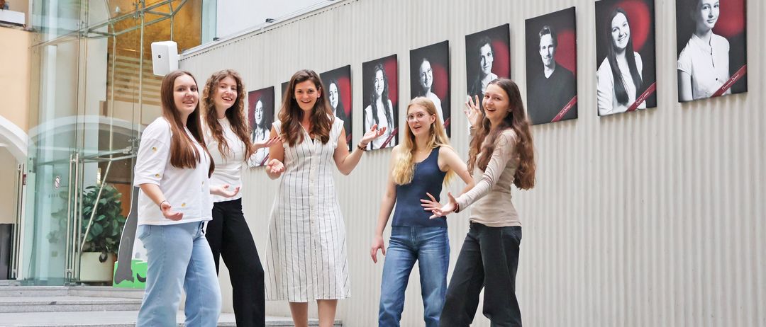 Fünf junge Frauen posieren vor einer Wand mit rot-thematischen Fotografien. Sie tragen lässige Kleidung und lächeln. Die Wand hat eine weiße und graue Metalloberfläche.