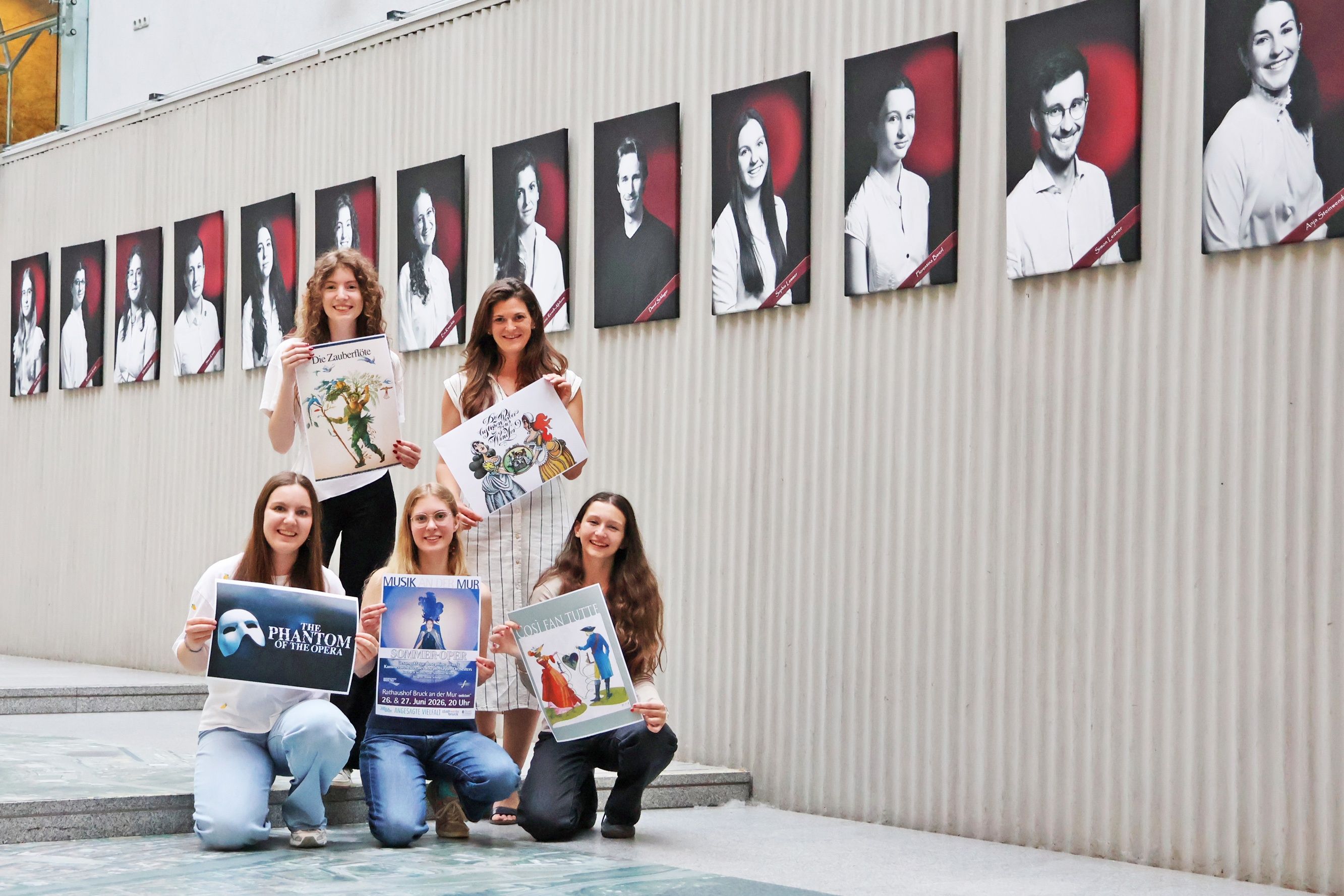 Fünf Frauen posieren mit Werbeplakaten vor einer Wand mit Porträts. Jedes Plakat zeigt einen anderen Event- oder Showtitel.