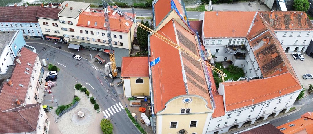 Eine Luftaufnahme von Gebäuden mit Baukränen auf ihren Dächern. In der Nähe sind geparkte Fahrzeuge, eine Straße, ein Fluss und Bäume.