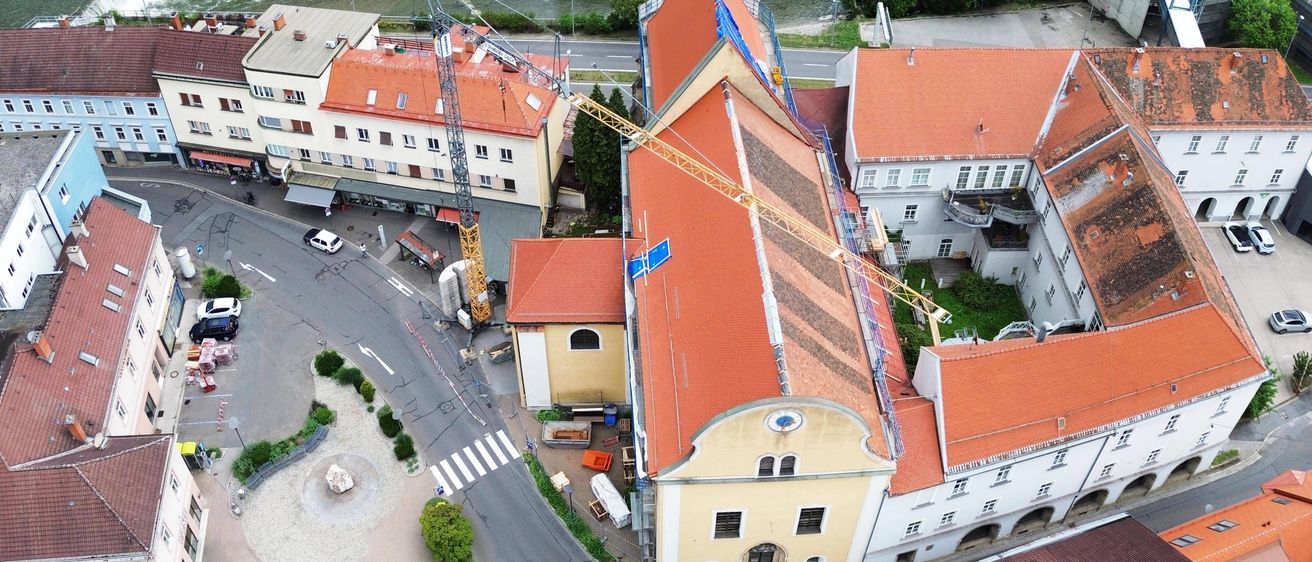 Eine Luftaufnahme von Gebäuden mit Baukränen auf ihren Dächern. In der Nähe sind geparkte Fahrzeuge, eine Straße, ein Fluss und Bäume.