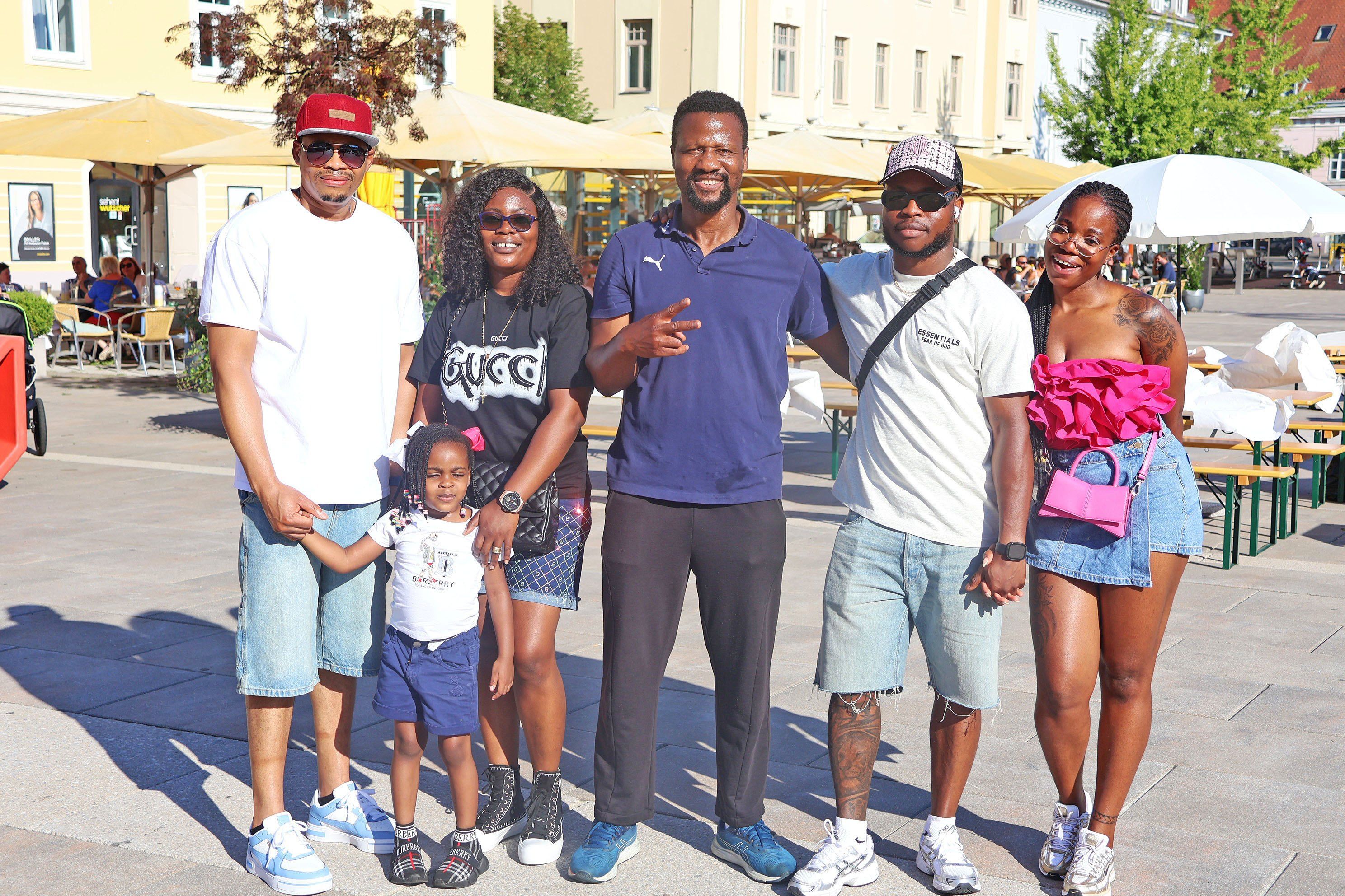Eine Familie mit fünf Personen, darunter ein junges Kind, posiert für ein Foto im Freien. Sie tragen Freizeitkleidung und stehen vor einem Gebäude mit Bäumen und Schirmen.