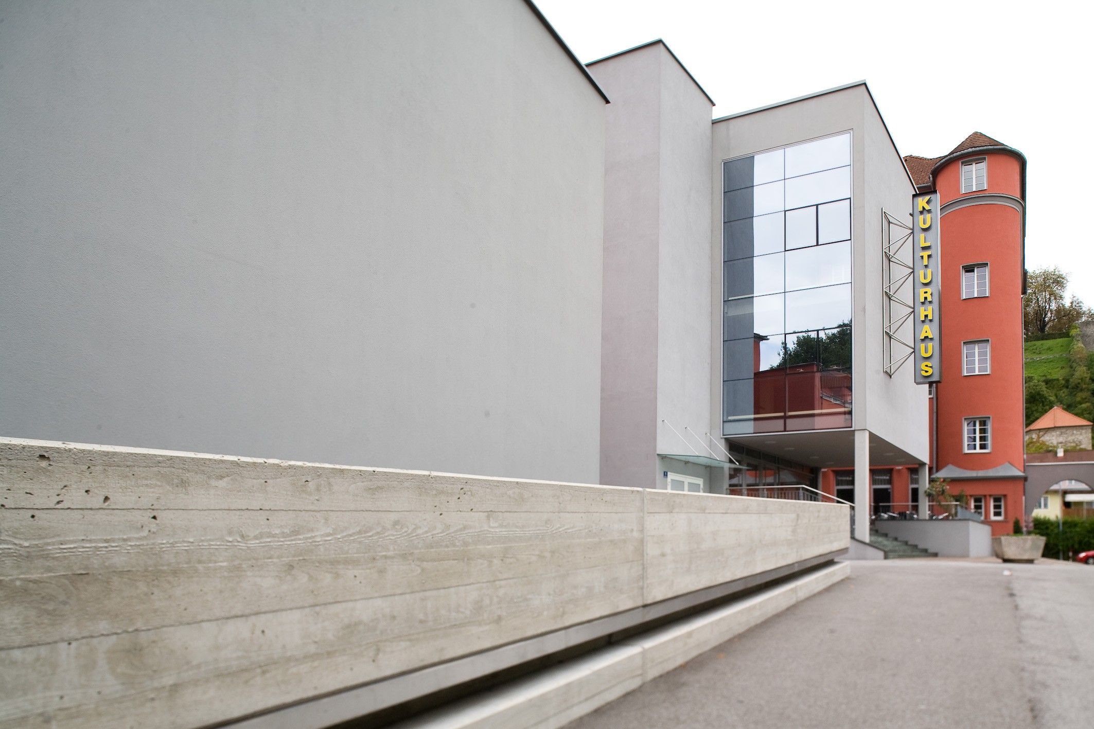 Ein modernes Gebäude mit einer Betonwand, Glasfenstern und einem Metallgeländer ist zu sehen. Die Wand ist grau mit einer strukturierten Oberfläche.