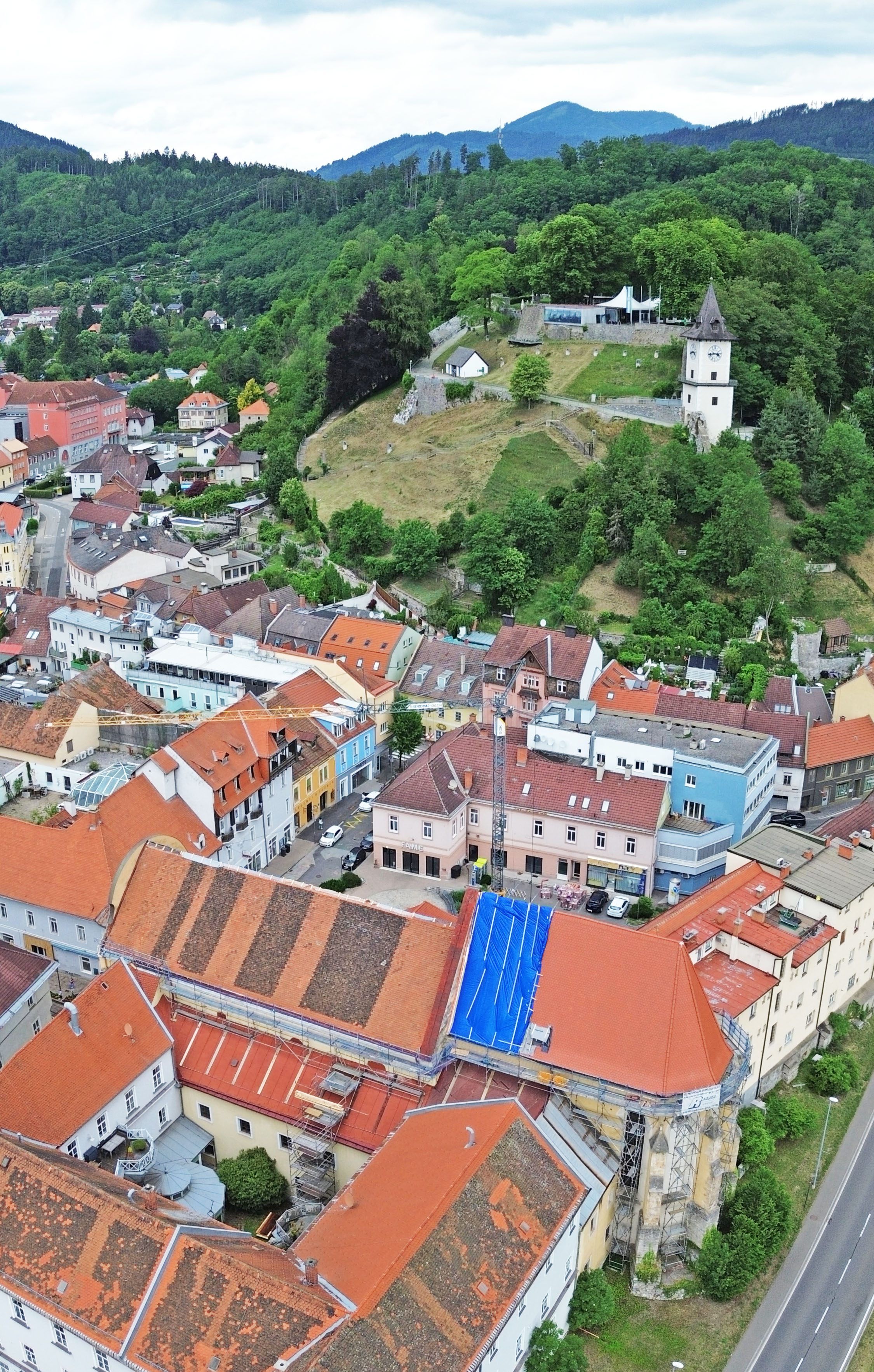 Eine Luftaufnahme einer Stadt mit mehreren Gebäuden, einige im Bau. Rote Dächer dominieren, mit einem auffälligen weißen Turm in der Ferne. Bäume und Grünflächen umgeben das Gebiet.