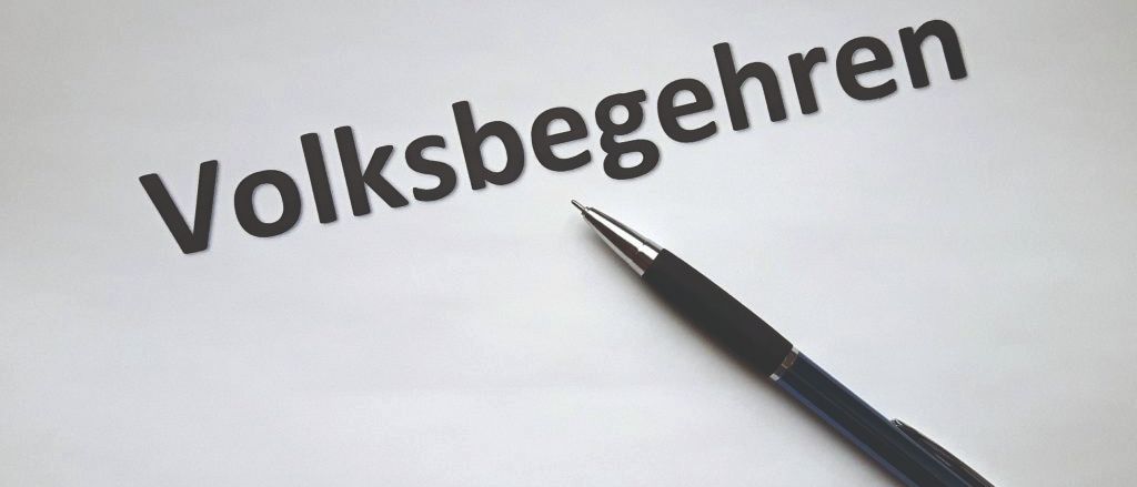 Ein schwarzer Stift liegt auf einem weißen Blatt Papier mit dem deutschen Wort 'Volksbegeehren' in schwarzer Tinte geschrieben.