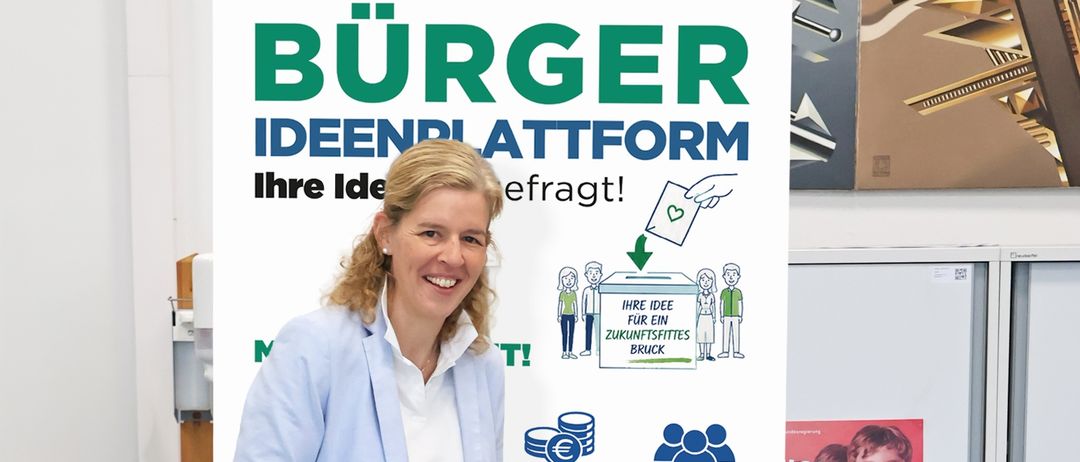Eine Frau steht neben einer Wahlurne, lächelt und hält ein Papier in der Hand, das sie in die Urne legt. Hinter ihr ist ein großes Plakat mit 'IDEENPLATTFORM' und 'Ihre Idee erfragt!' geschrieben.