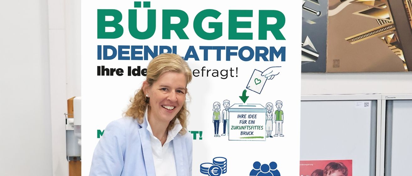 Eine Frau steht neben einer Wahlurne, lächelt und hält ein Papier in der Hand, das sie in die Urne legt. Hinter ihr ist ein großes Plakat mit 'IDEENPLATTFORM' und 'Ihre Idee erfragt!' geschrieben.