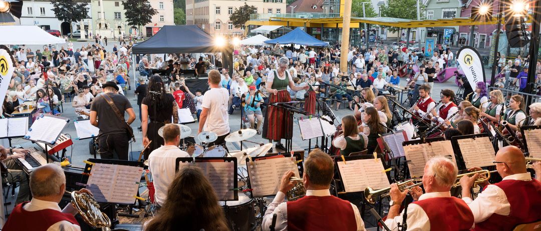 Ein Outdoor-Musikaufführung mit einem Publikum auf einem Stadtplatz, Musiker spielen Instrumente und eine Frau singt. Gebäude und Berge im Hintergrund.