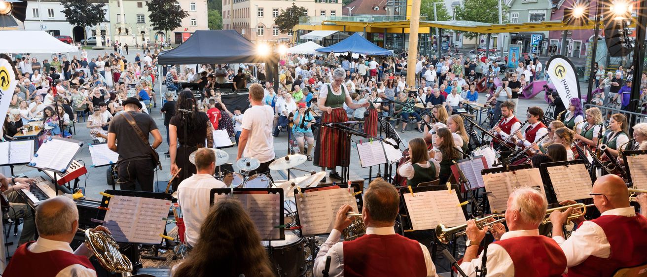 Ein Outdoor-Musikaufführung mit einem Publikum auf einem Stadtplatz, Musiker spielen Instrumente und eine Frau singt. Gebäude und Berge im Hintergrund.