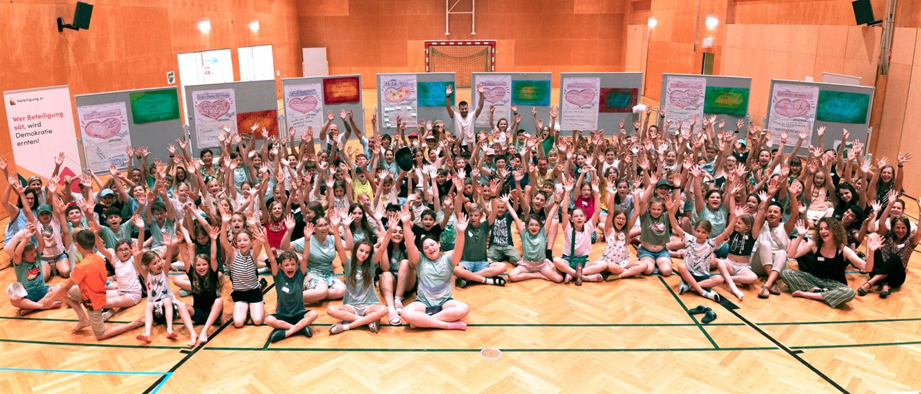 Eine Gruppe von Kindern und Erwachsenen posiert für ein Foto in einer Turnhalle mit Holzboden und Wänden mit Kunst.