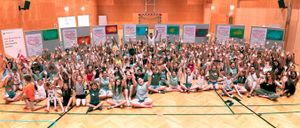 Eine Gruppe von Kindern und Erwachsenen posiert für ein Foto in einer Turnhalle mit Holzboden und Wänden mit Kunst.
