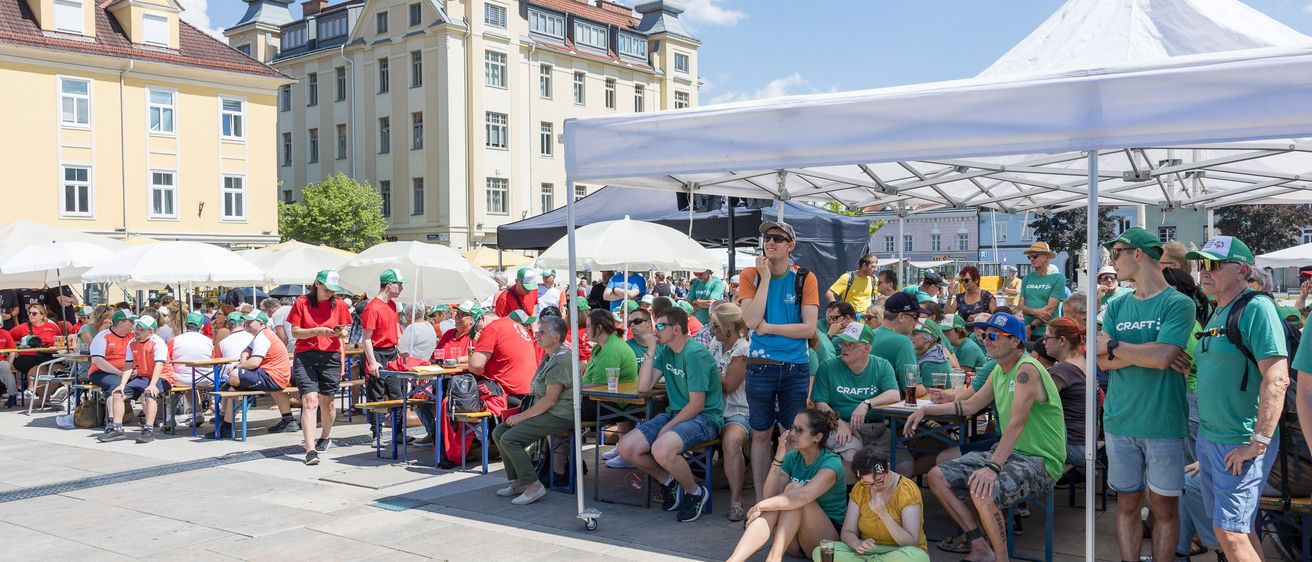 Viele Menschen in grünen T-Shirts sitzen unter einem Zelt auf einer Straße und unterhalten sich, mit einem Gebäude im Hintergrund.