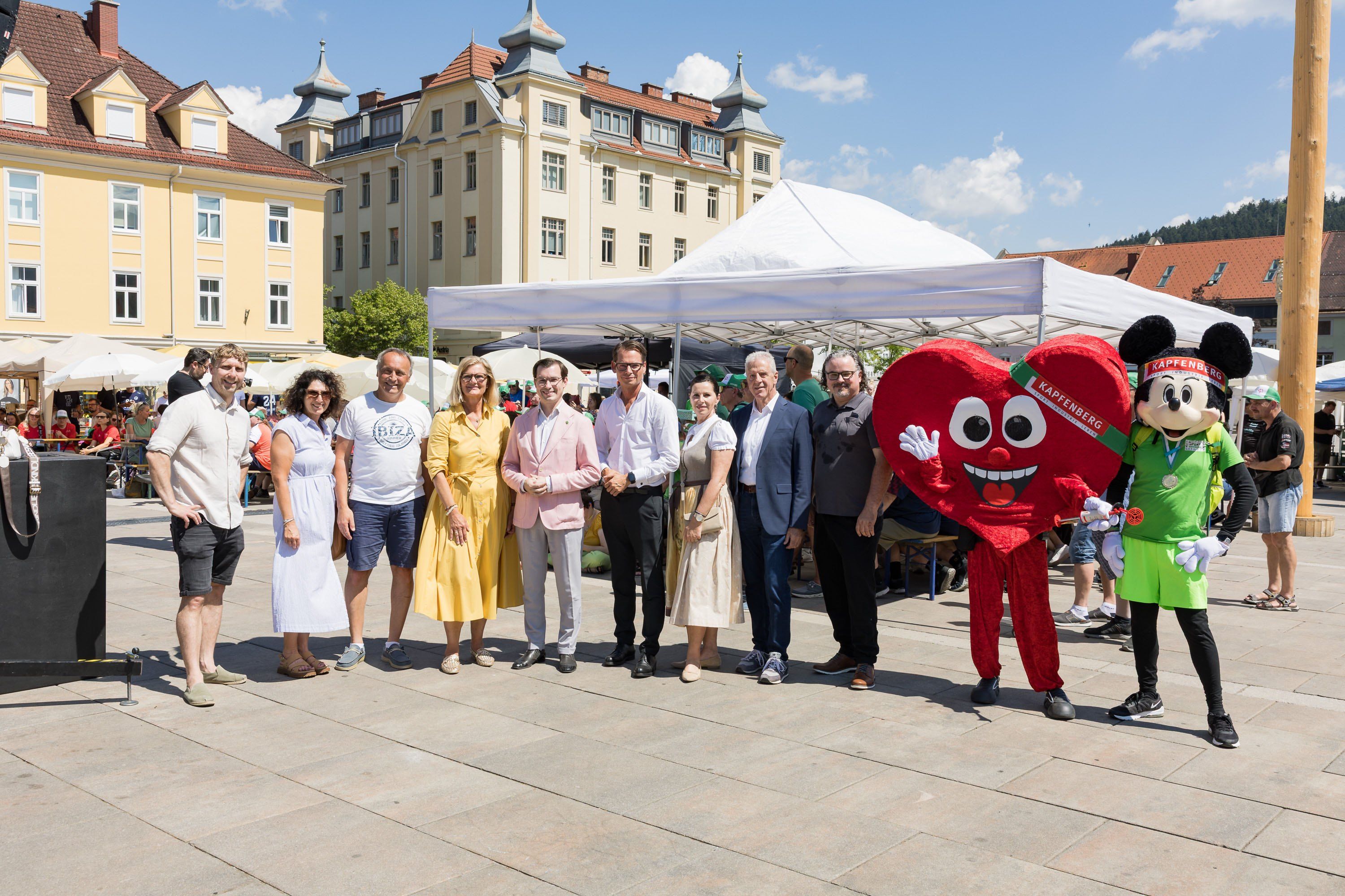 Eine Gruppe von Menschen steht zusammen auf einem Platz, posiert für ein Foto mit einem roten Herz-Maskottchen, vor einem weißen Zelt und Gebäuden.