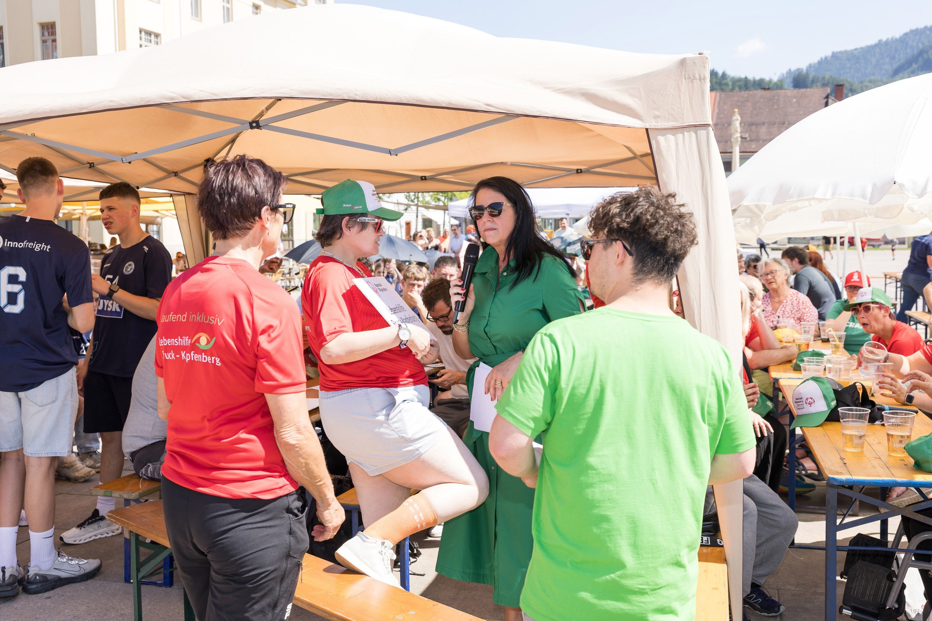 Eine Frau in Grün spricht in ein Mikrofon unter einem Zelt, während andere zuhören. Ein Mann in einem roten Shirt steht hinter ihr, und ein Mann in Grün steht ihr gegenüber.