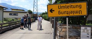 Eine Gruppe von Menschen steht auf einem Bürgersteig mit einem gelben Schild, das nach links zeigt. Das Schild sagt 'Umleitung'. Eine Steinmauer befindet sich rechts. Bäume, Häuser und ein Turm sind im Hintergrund.