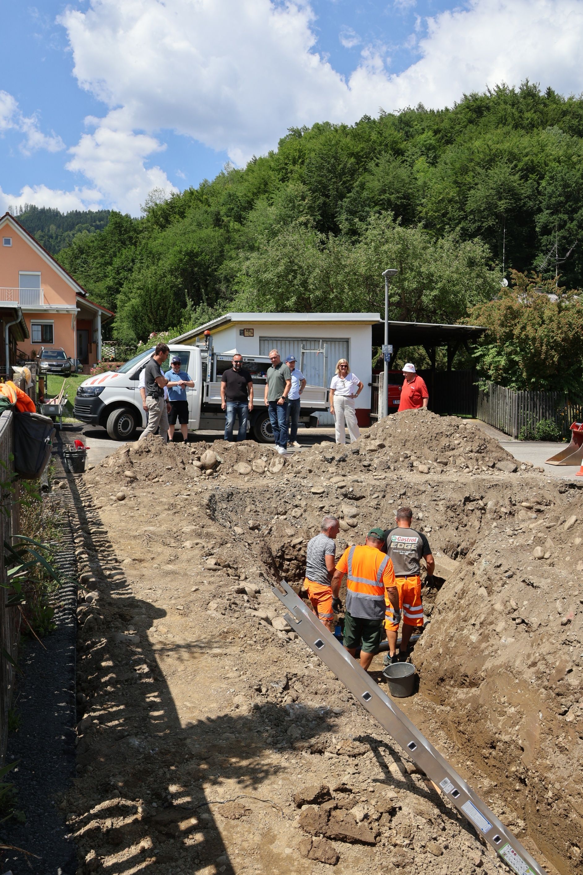 Eine Gruppe Arbeiter gräbt eine Grube vor einem Haus. Ein Lieferwagen ist in der Nähe geparkt. Bäume und Berge sind im Hintergrund.