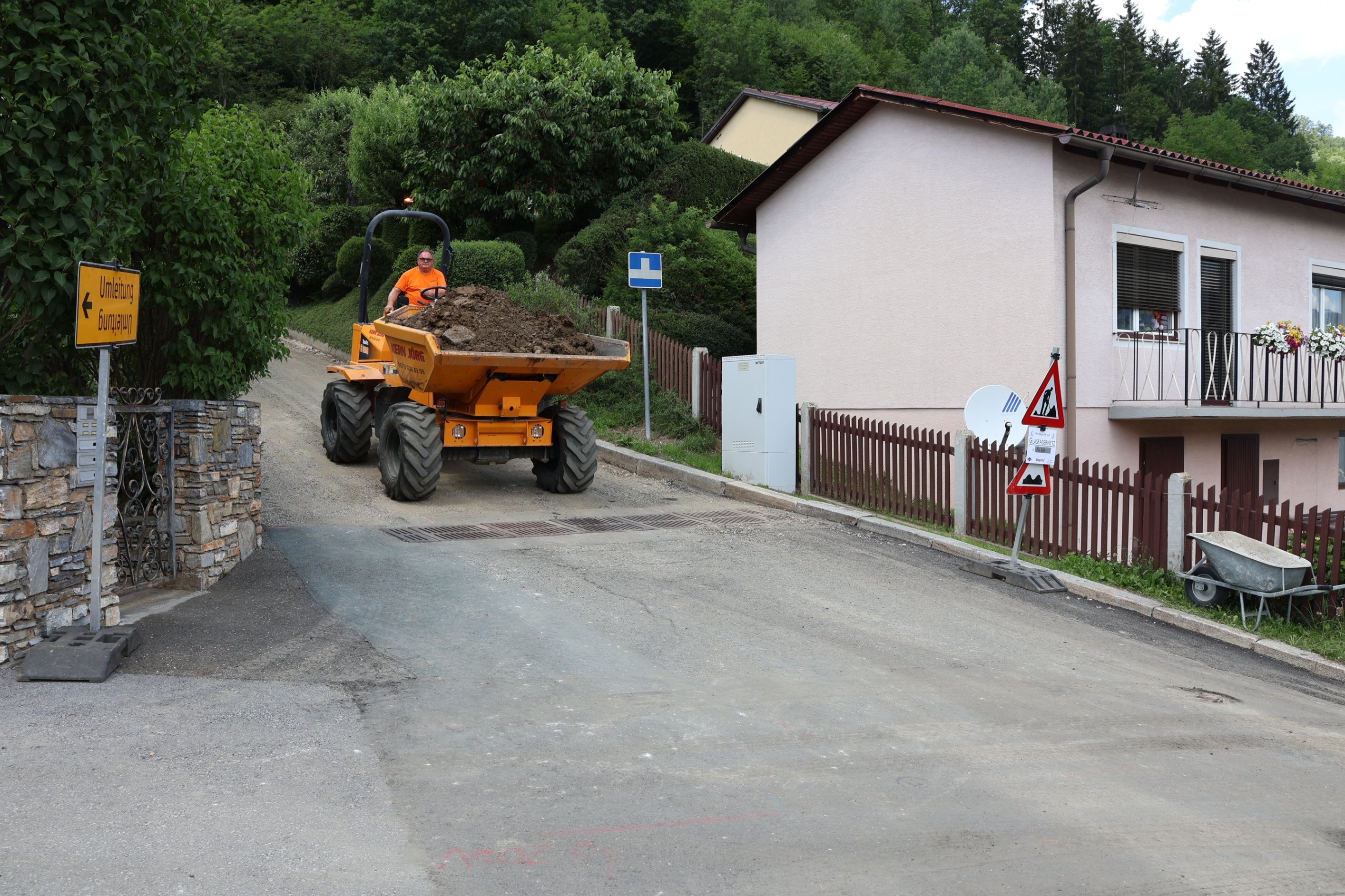 Ein Mann fährt einen gelben Traktor mit voller Erde auf einer Einfahrt, mit einem Haus und einem Zaun im Hintergrund.