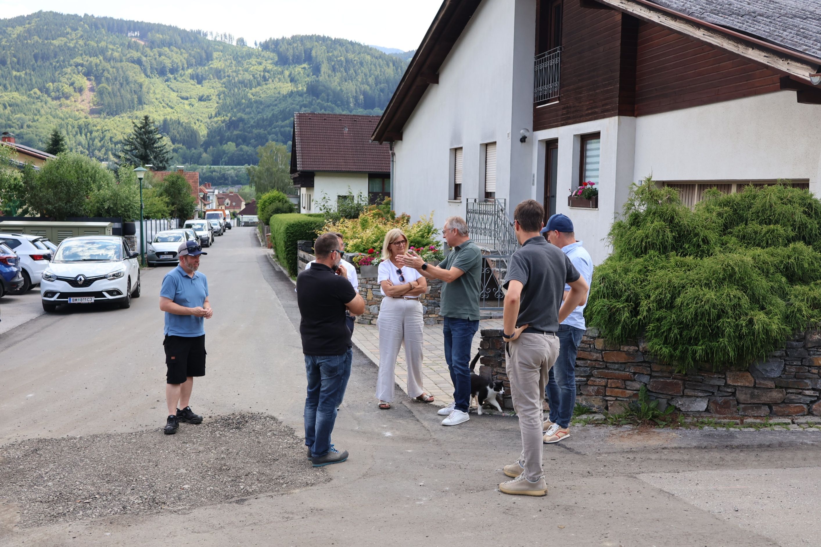 Eine Gruppe von Menschen steht draußen und führt ein Gespräch. Ein Mann hält eine Kamera. In der Nähe sind eine Frau mit Brille und ein Hund anwesend. Die Szene umfasst Häuser, Grünflächen und geparkte Autos. Berge sind im Hintergrund sichtbar.