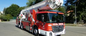 Ein roter Mercedes-Benz Feuerwehrwagen mit ausgefahrenem Drehleiter steht auf einem Parkplatz. Vorne steht das Wort 'FEUERWEHR'.