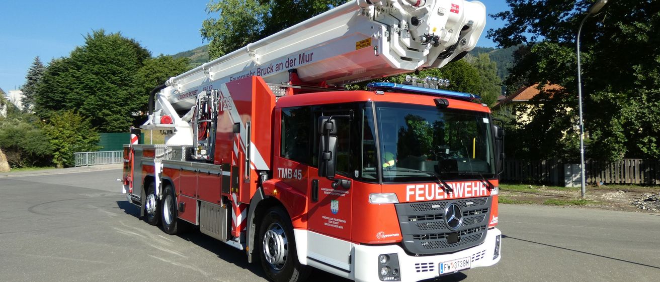 Ein roter Mercedes-Benz Feuerwehrwagen mit ausgefahrenem Drehleiter steht auf einem Parkplatz. Vorne steht das Wort 'FEUERWEHR'.