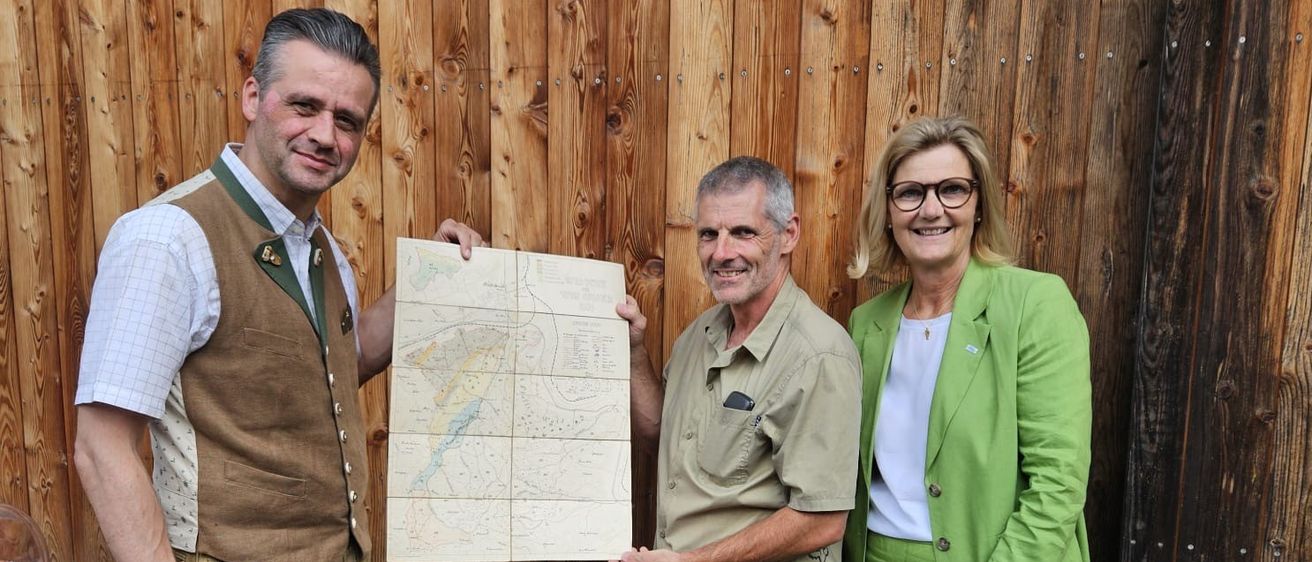Drei Personen stehen vor einer Holzwand und halten eine Karte in der Hand. Der Mann links trägt traditionelle bayerische Kleidung, während die anderen zwei Freizeitkleidung tragen.