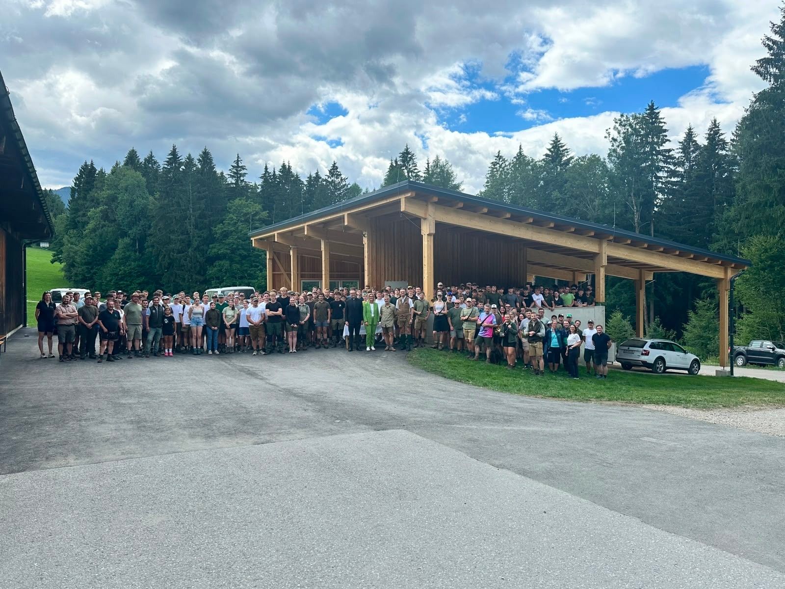 Eine große Gruppe von Menschen in Outdoor-Kleidung steht vor einem hölzernen Pavillon mit einem Waldhintergrund.