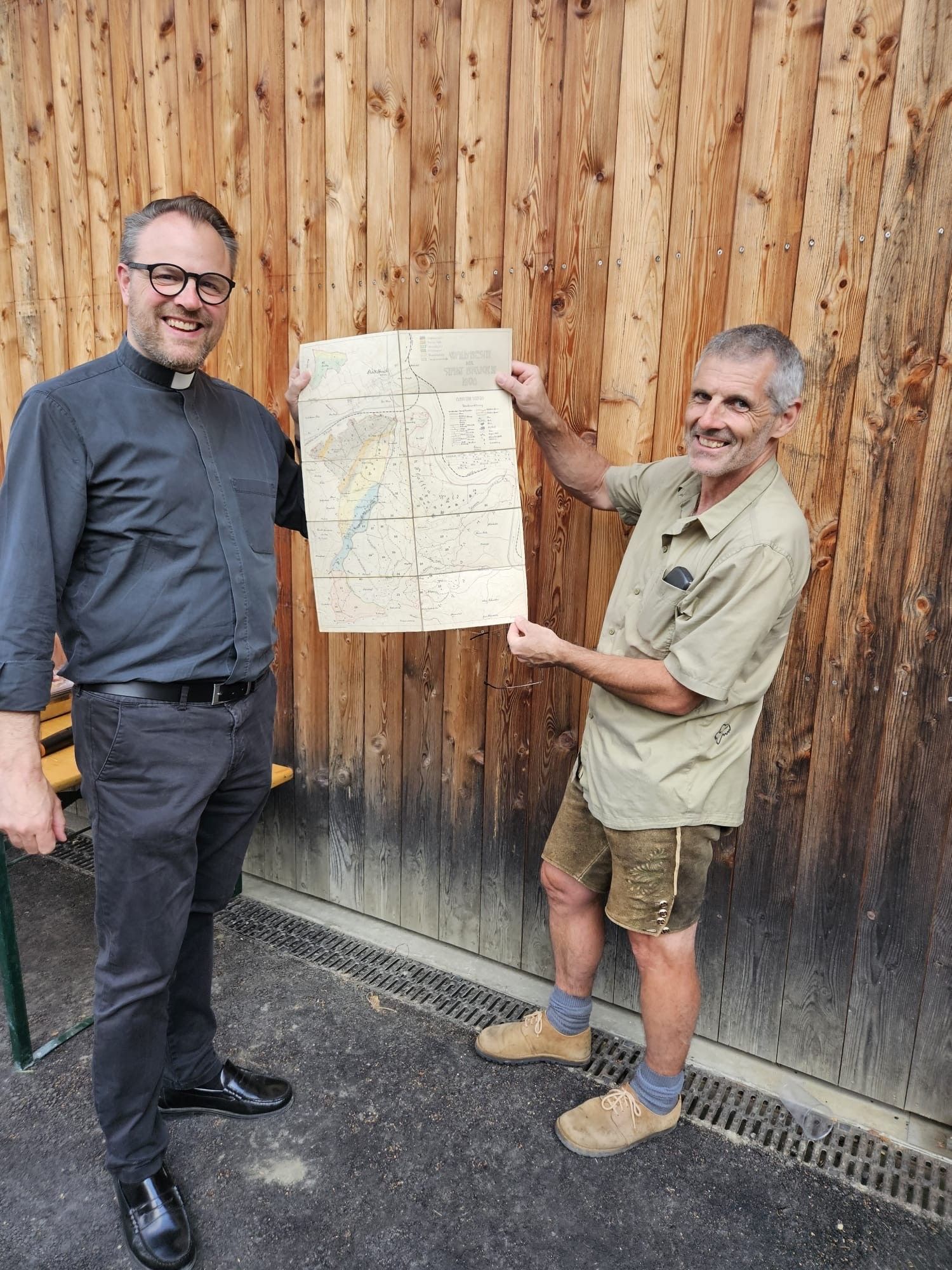 Zwei Männer stehen vor einer Holzwand, einer hält eine Karte in der Hand und beide lächeln. Der Mann auf der linken Seite trägt ein schwarzes Hemd mit Kragen und eine Brille, während der Mann auf der rechten Seite ein grünes Hemd und Shorts trägt.
