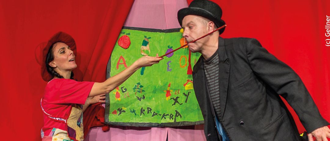 Ein Werbeposter für eine Theaterveranstaltung. Eine Frau und ein Mann stehen mit einem bemalten Schild. Die Veranstaltung heißt "Die kleine Hexe & der kleine Rabe" und ist für den 1. März 2026 geplant.