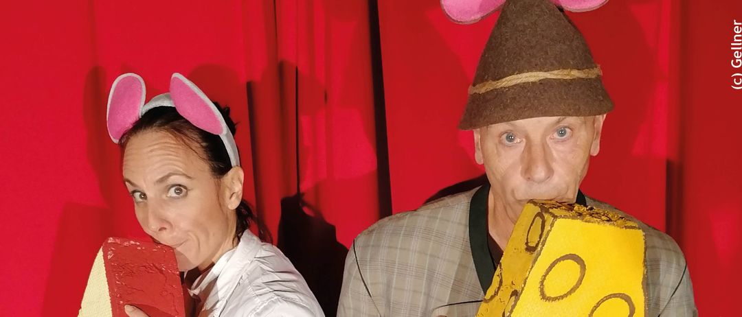 Plakat für eine Theateraufführung mit zwei Erwachsenen in Kostümen. Die Frau hält ein rotes Objekt und der Mann ein gelb-käseförmiges Objekt. Das Plakat enthält Datum und Uhrzeit der Veranstaltung.