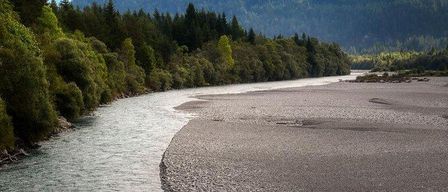 Ein Fluss fließt durch ein felsiges Gelände mit üppigen Bäumen am linken Ufer und einem entfernten Bergrücken.