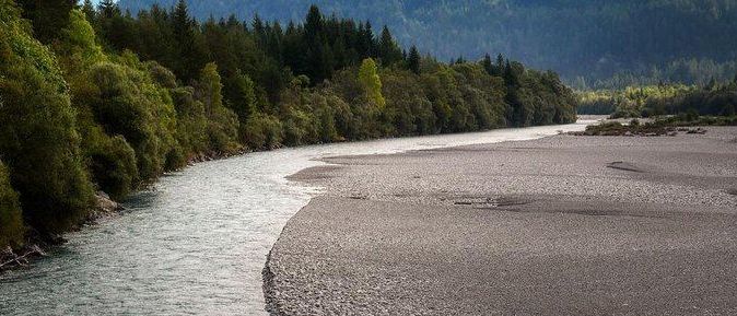Ein Fluss fließt durch ein felsiges Gelände mit üppigen Bäumen am linken Ufer und einem entfernten Bergrücken.