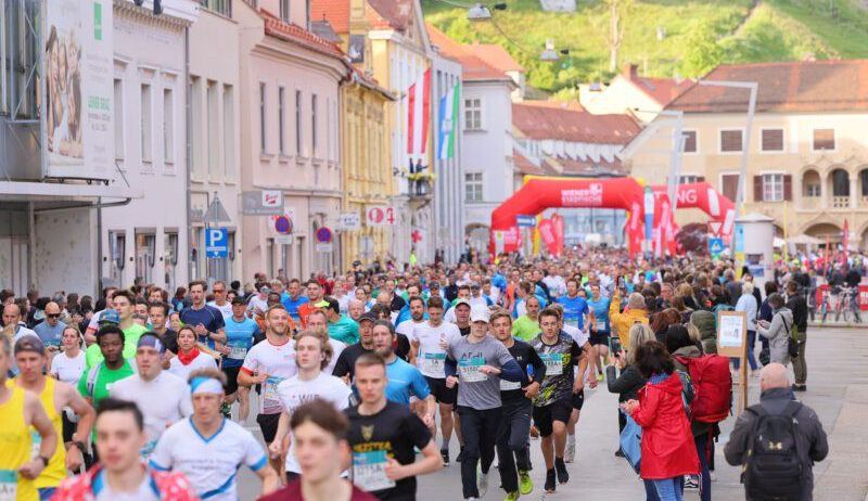 Eine Menge Läufer bei einem Marathon auf einer Stadtstraße mit einem roten Bogen im Hintergrund. Zuschauer säumen den Bürgersteig.