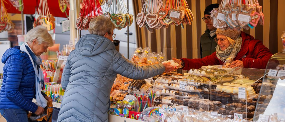 Zwei Frauen an einem Lebensmittelstand mit verschiedenen Süßigkeiten, eine Frau gibt dem Verkäufer Geld. Oben hängen Bonbons und Kekse.