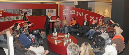 Eine Gruppe von Menschen sitzt in Stühlen um einen Tisch in einem Raum mit roten Wänden. Eine Frau steht am Tisch und leitet möglicherweise eine Diskussion.