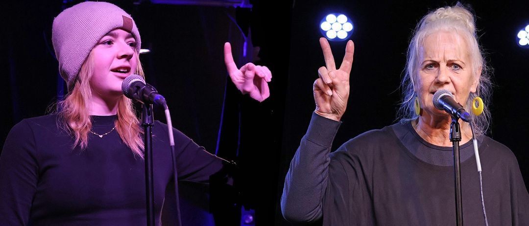 Zwei Frauen auf der Bühne, eine singt in ein Mikrofon und die andere macht ein Friedenszeichen. Beide tragen dunkle Oberteile mit langen Ärmeln.