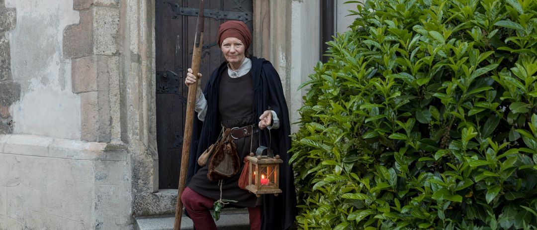 Eine Frau in mittelalterlicher Kleidung mit einer Laterne steht auf den Stufen eines alten Gebäudes.