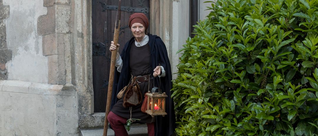 Eine Frau in mittelalterlicher Kleidung hält eine Lanze und eine Laterne vor einem Gebäude mit einer Holztür.