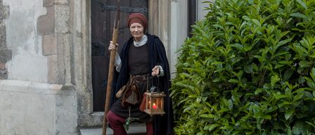 Eine Frau in mittelalterlicher Kleidung hält eine Lanze und eine Laterne vor einem Gebäude mit einer Holztür.
