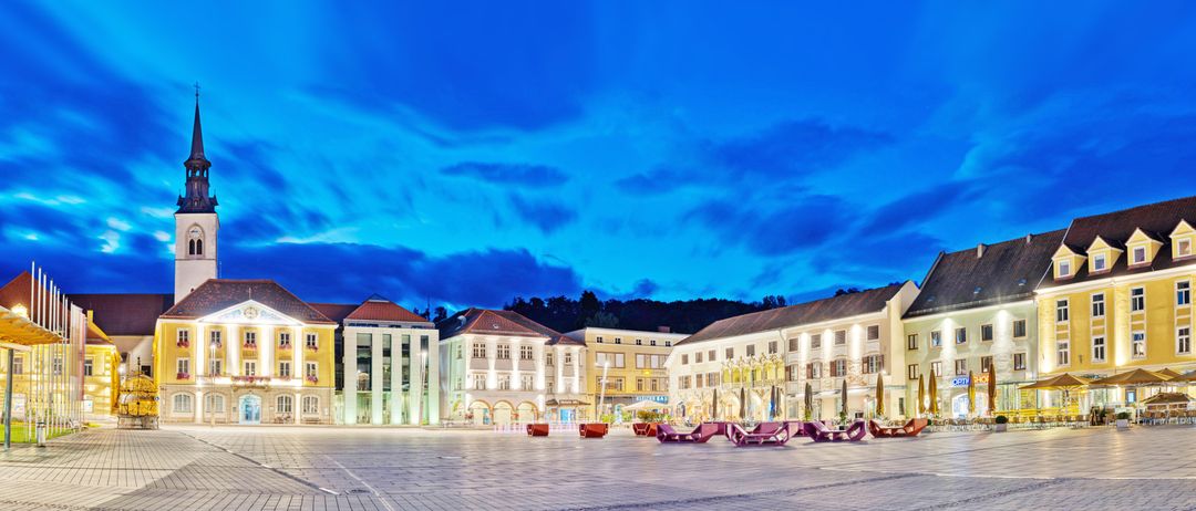 Ein Stadtplatz mit beleuchteten Gebäuden unter einem blauen Himmel mit Wolken und einer Reihe lila Stühle vor den Gebäuden.