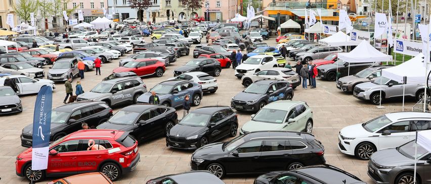 Viele Autos sind auf einem Stadtplatz geparkt. Es gibt Leute, die herumlaufen. Die Autos sind in verschiedenen Farben und Modellen. In der Nähe gibt es einige Zelte und Flaggen.