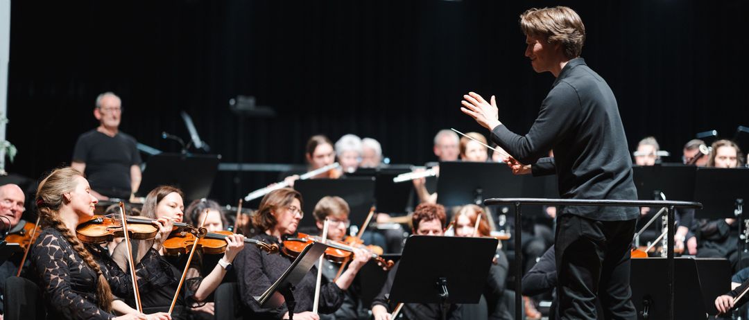 Ein Mann dirigiert ein Orchester auf der Bühne, mit Geigern vor ihm. Hinter ihm spielen Musiker verschiedene Instrumente unter Scheinwerfern.