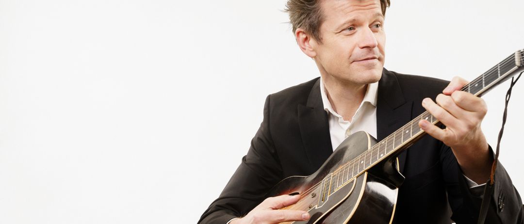 Ein Mann in einem Anzug spielt eine Gitarre und schaut in die Kamera. Die Gitarre hat einen goldbraunen Korpus und einen schwarzen Hals.